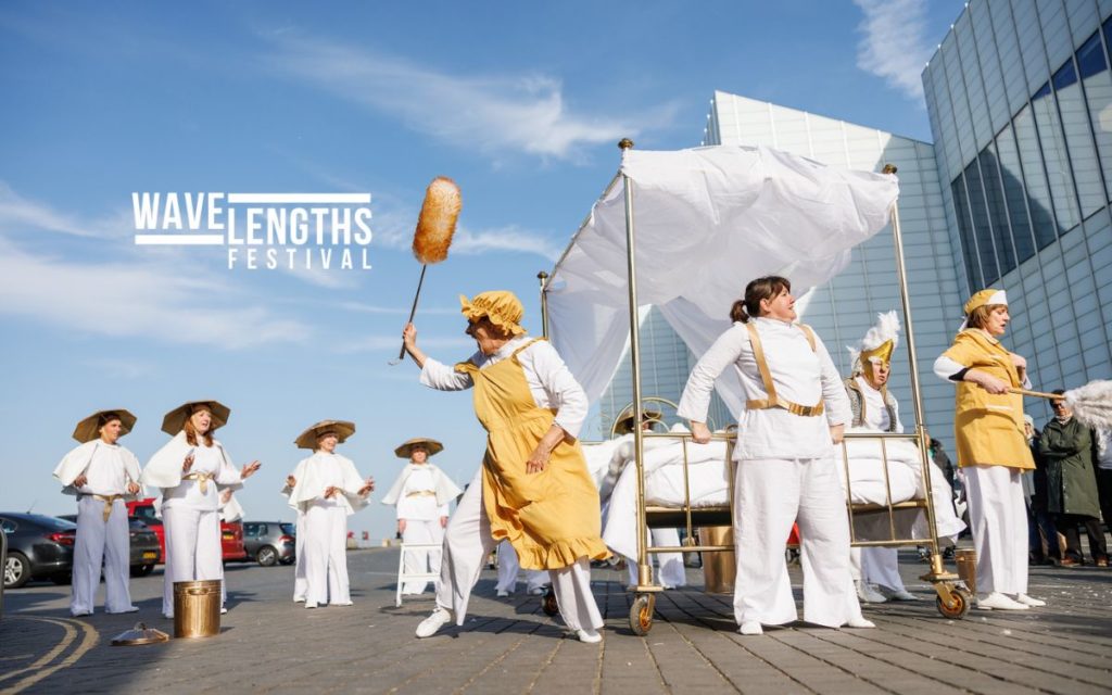 A group of performance in white and gold costumes perform with a golden bed on wheels outdoors. The logo 'Wavelengths Festival' overlays the image.