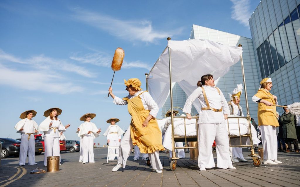 A group of performance in white and gold costumes perform with a golden bed on wheels outdoors.