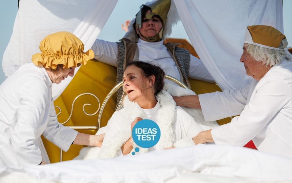 Three women in white and gold costume attend to another figure lying in a golden bed with white linens. The blue Ideas Test logo overlays the image.