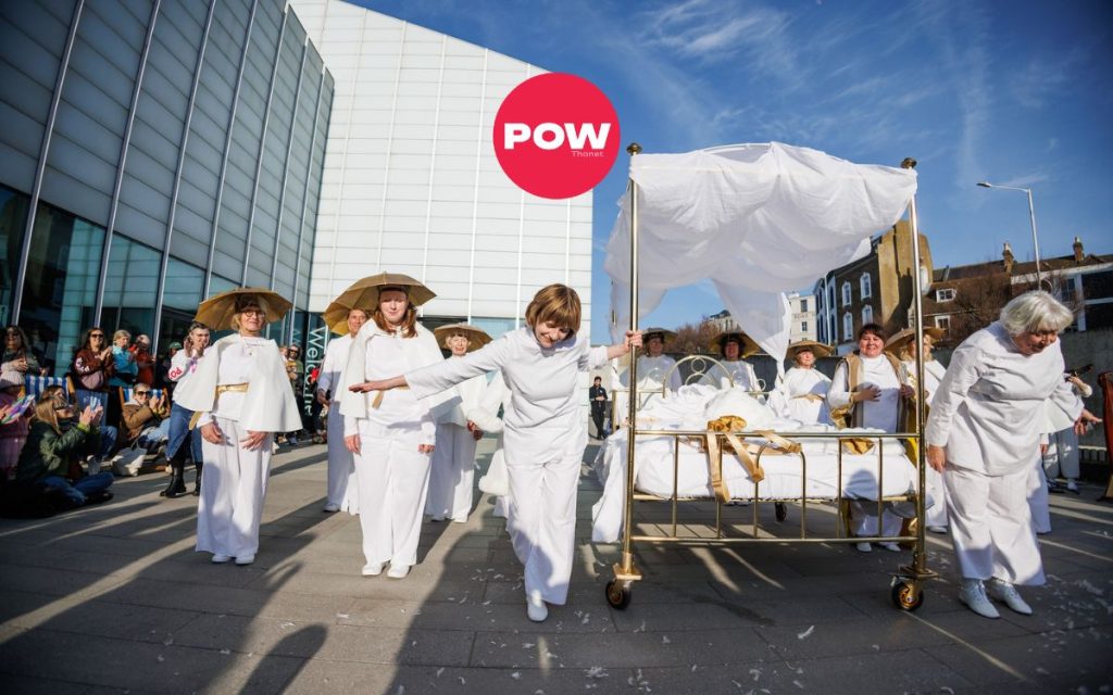A group of performers in white costumes surround a golden bed with white linens. The red circular logo "POW Thanet" logo overlays the image.