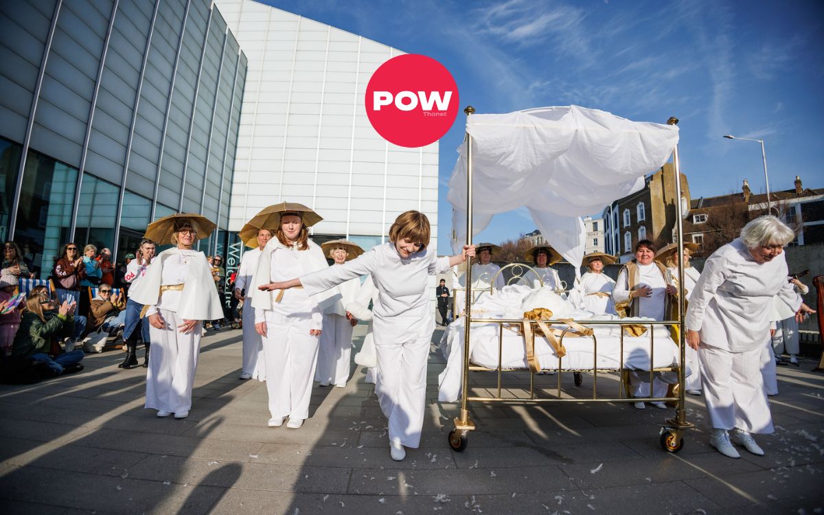 A group of performers in white costumes surround a golden bed with white linens. The red circular logo "POW Thanet" logo overlays the image.