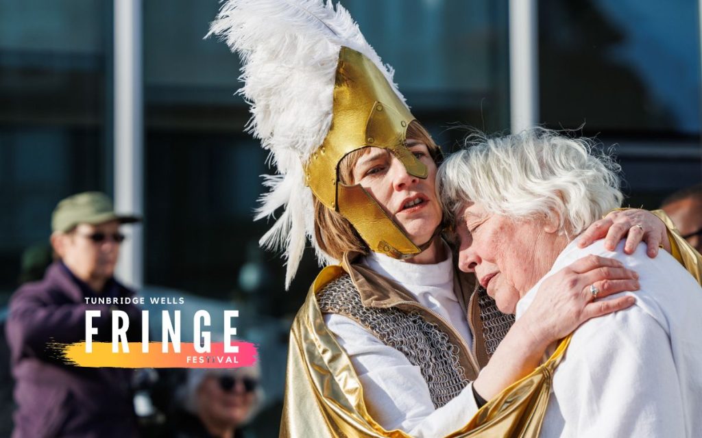 A woman in a golden helmet and feathered headpiece embraces another person with white hair. The Tunbridge Wells Fringe Festival logo overlays the image.