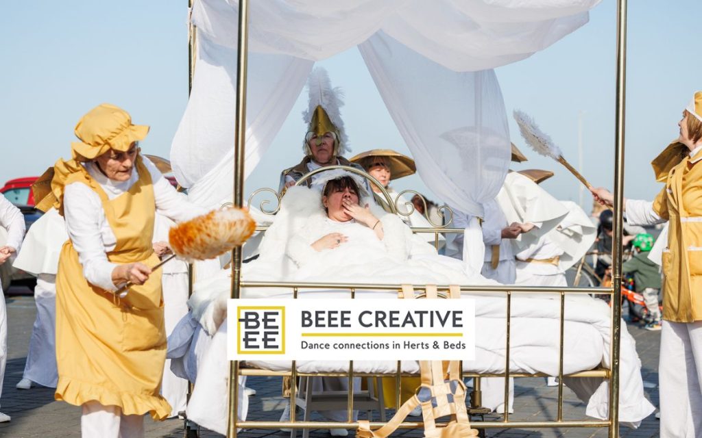 A woman lies in a golden bed surrounded by others in white and gold costume, some holding feather dusters. The white and yellow Beee Creative logo overlays the image.