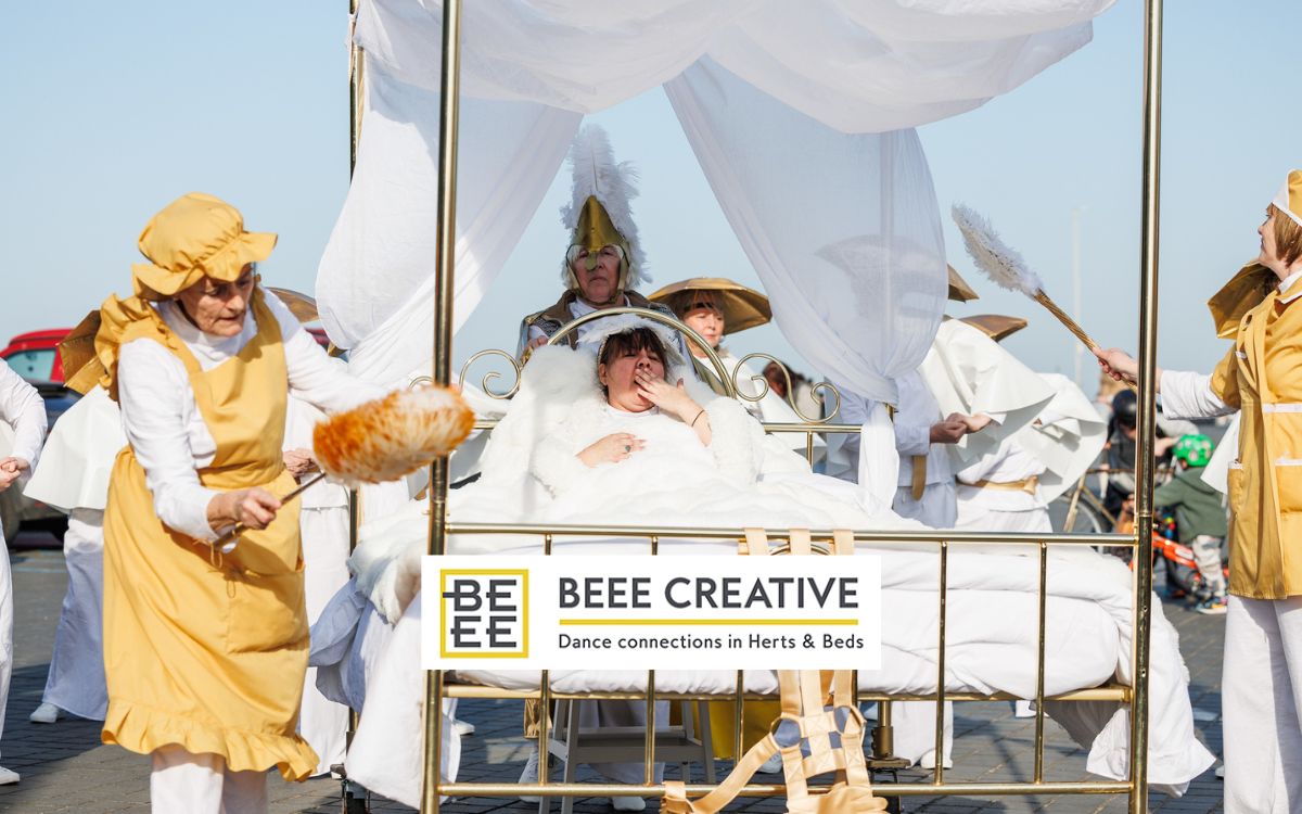 A woman lies in a golden bed surrounded by others in white and gold costume, some holding feather dusters. The white and yellow Beee Creative logo overlays the image.
