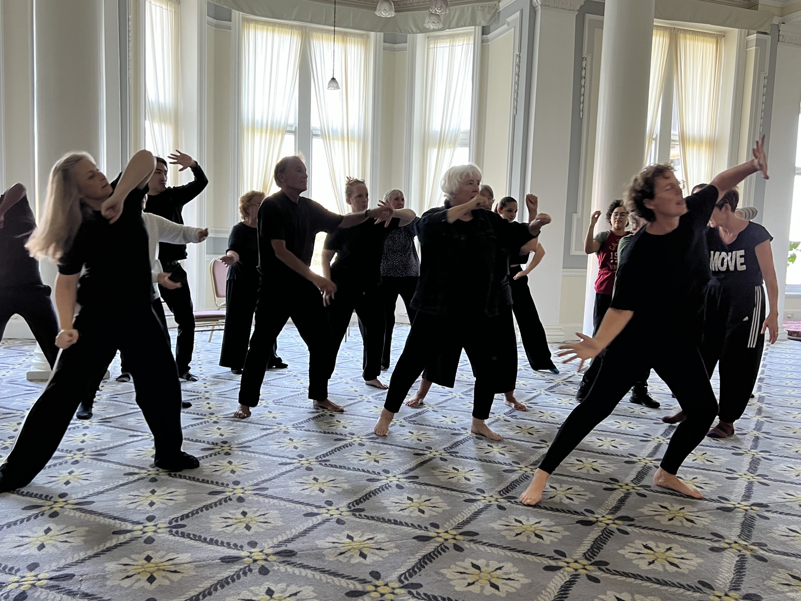 A group of performers in black clothing participate in a dance in a bright, airy room with large windows and a patterned carpet.