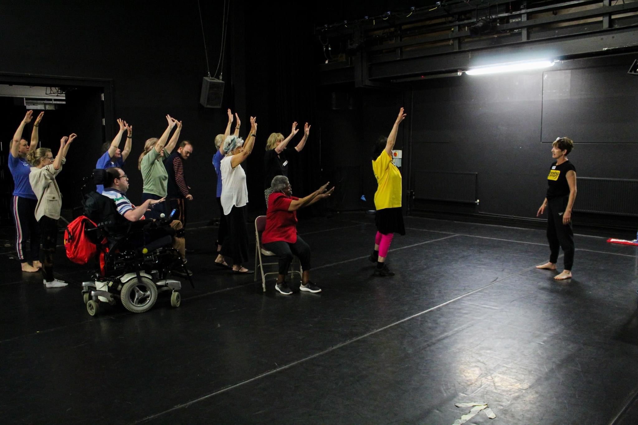 A woman with short hair in a Moving Memory T-shirt stands in front of a group of people, some standing, some in wheelchairs lifting their hands in the air.