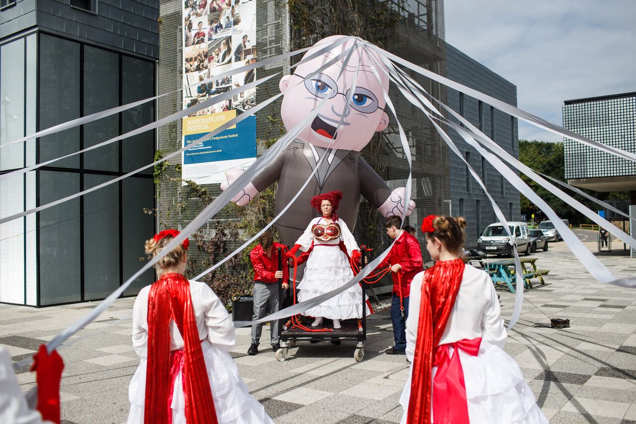 A giant inflatable man wearing glasses and dressed in a suit is attached to giant maypole ribbons as women in white dresses and red head dresses hold the giant ribbons. A women with a white dress and red wig stands defiantly in front of the giant inflatable puppet.