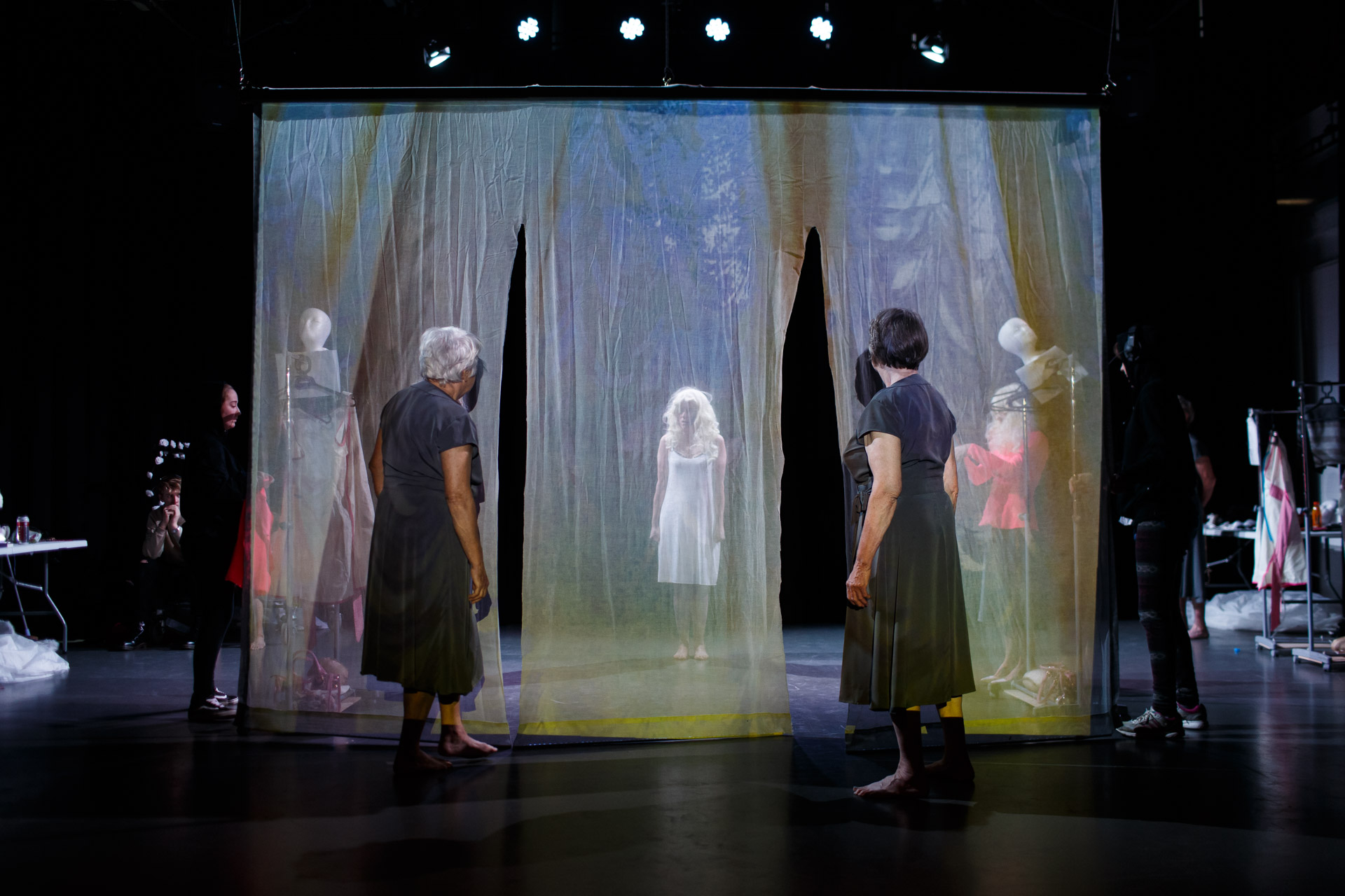 Two women in grey dresses stand with their backs to the audience and look through white gauze where a woman in a white slip dress and blonde wig stands.