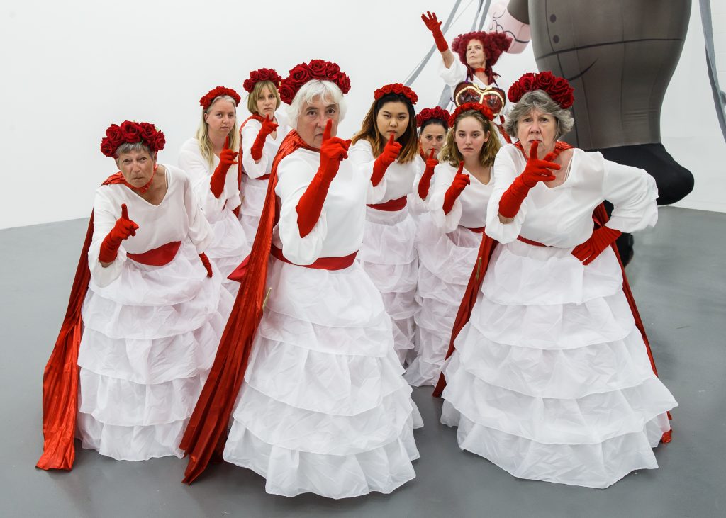 9 women dressed in frilly white dresses with red rose head dresses, scarves and gloves stare solemnly at the camera and wag their fingers.
