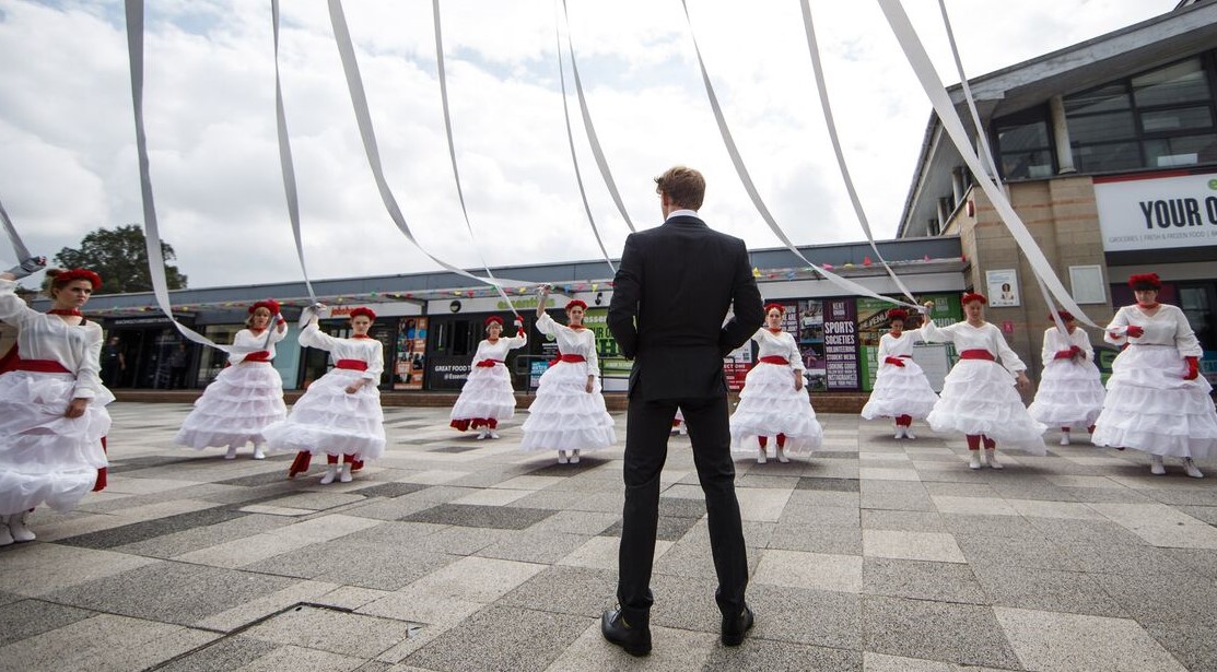 Women in white dresses with red head dresses hold on to giant maypole ribbons as a man in a black suit looks on.