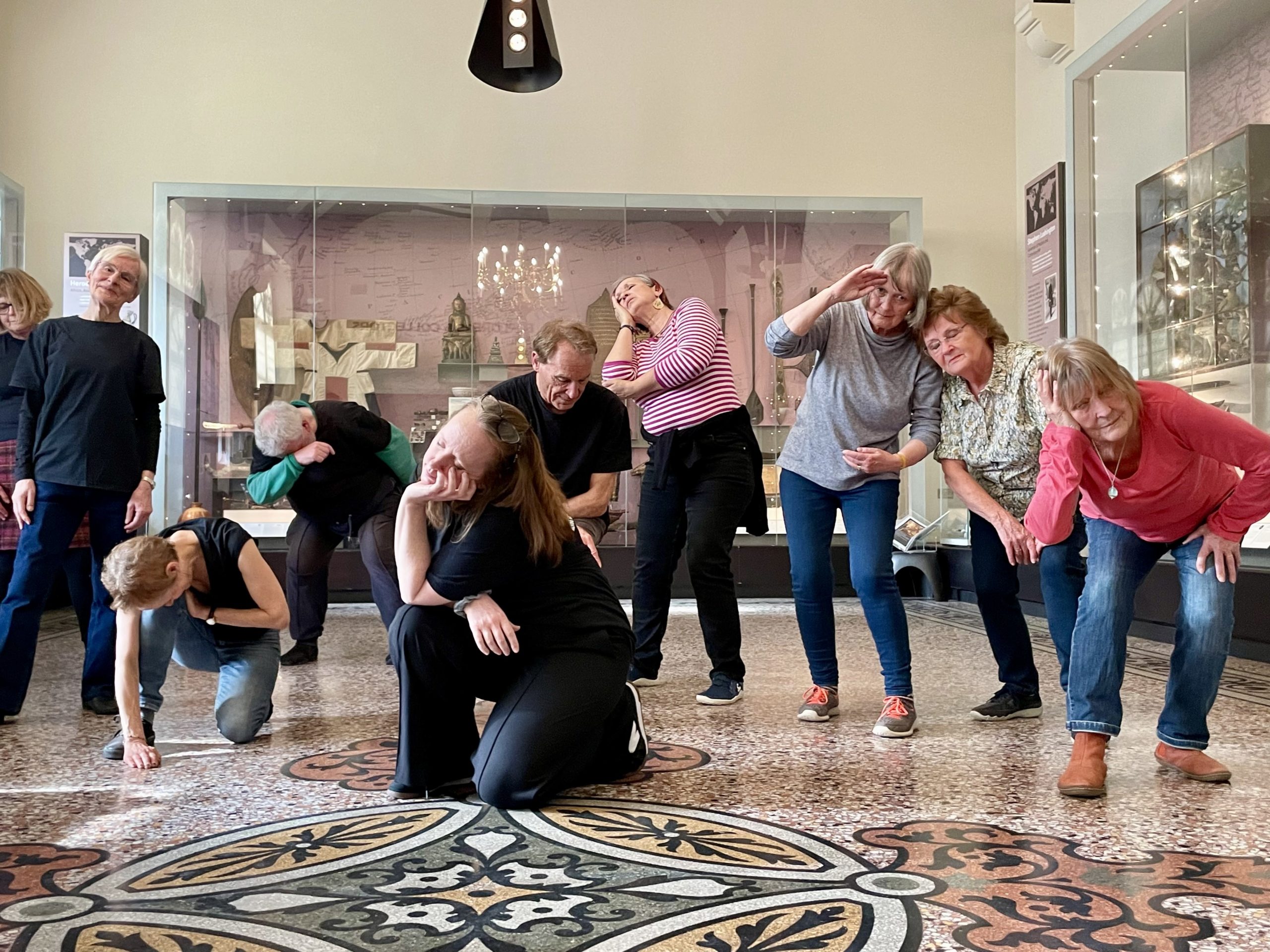 A group of people are performing in a museum setting. They are in various poses, some kneeling, some standing, in front of a glass display case and on a patterned floor.