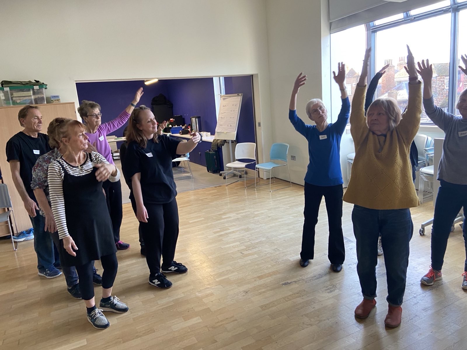A group of adults are standing in a bright room with a wooden floor, participating in a movement class. Some have their arms extended, while others have their hands near their chests.