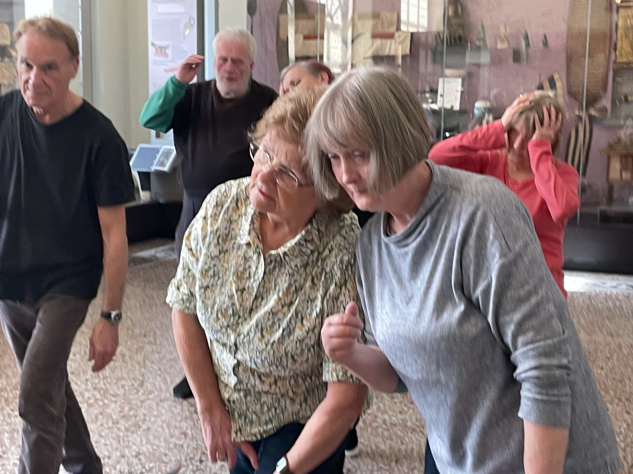 A group of people in a museum are participating in a movement class. Some have their hands near their faces. The floor is patterned.