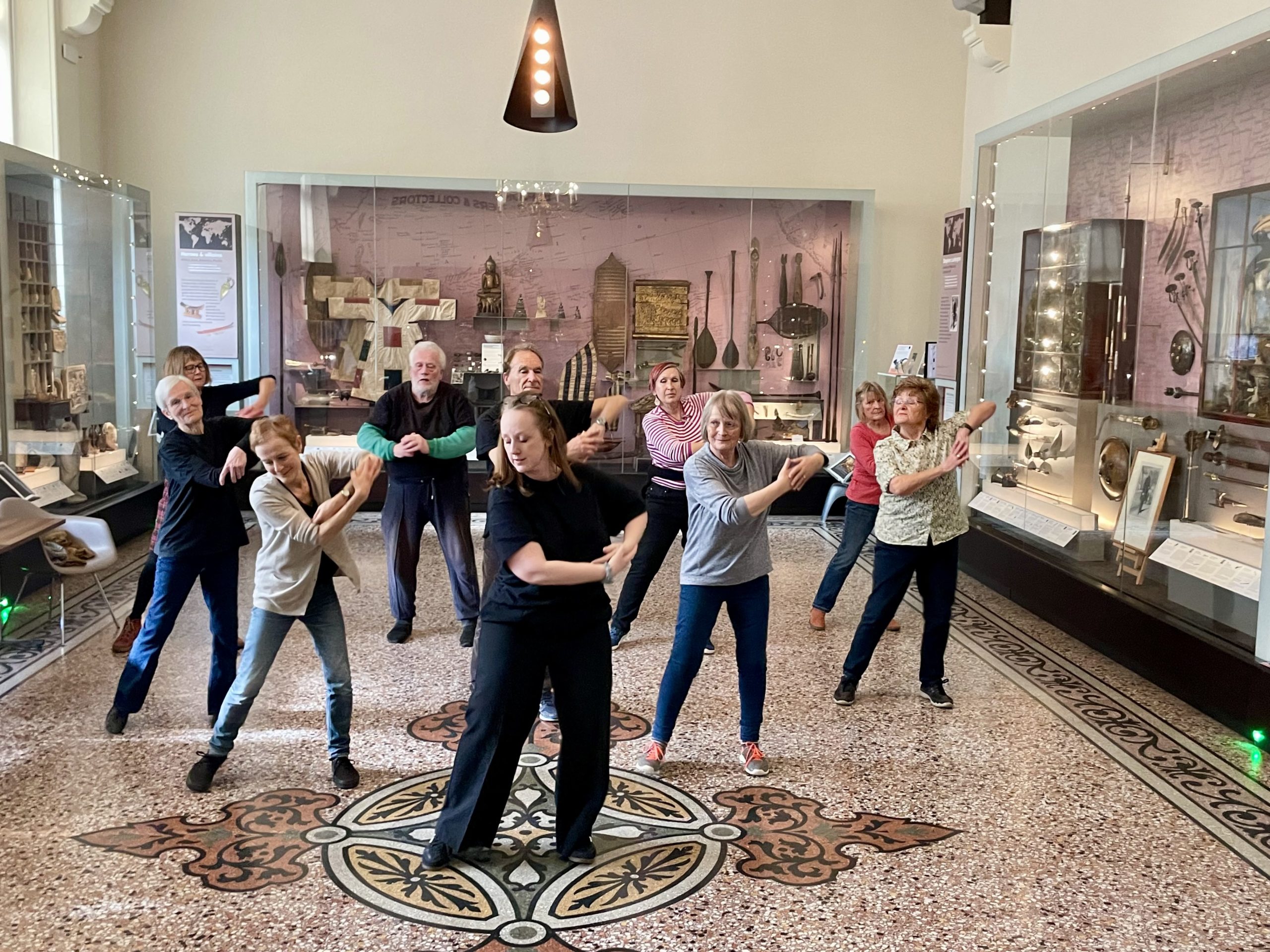 A group of adults are in a museum space with display cases, engaging in a movement activity. Their holding their own hands in a rocking motion.
