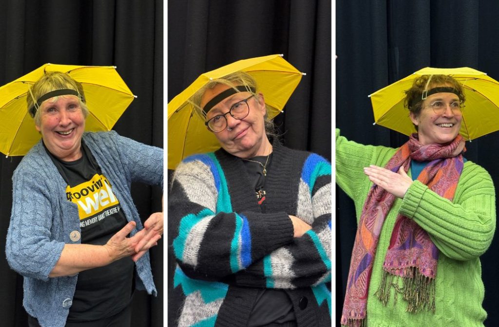 Three smiling women wear yellow umbrella hats against a dark backdrop. The person on the left has short brown hair and a blue cardigan, the center person has gray hair and glasses with crossed arms, and the rightmost person has curly brown hair and a green sweater with a colorful scarf.