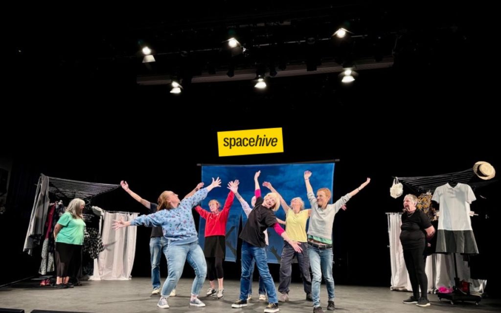 A group of people on a stage performing or rehearsing under overhead lights, with some posing with raised arms. Clothing racks with garments are visible on both sides of the stage. A yellow 'spacehive' logo is overlaid above the group.