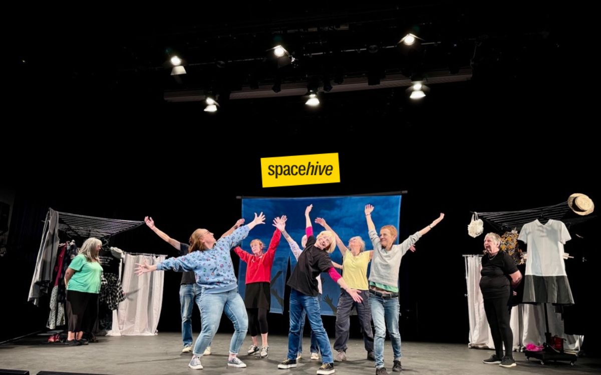 A group of people on a stage performing or rehearsing under overhead lights, with some posing with raised arms. Clothing racks with garments are visible on both sides of the stage. A yellow 'spacehive' logo is overlaid above the group.