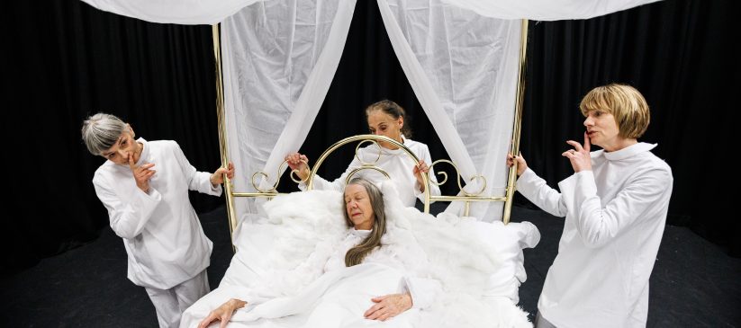 Four women in white clothes. One is lying on a white bed with a canopy. The other three are standing around the bed. The background is black.