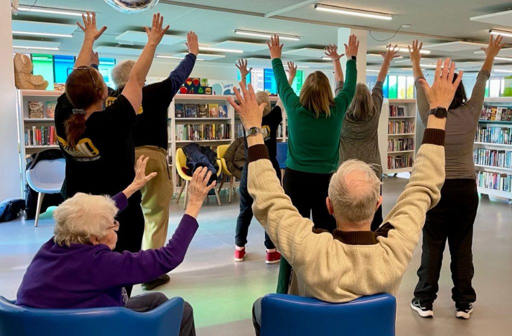 A group of adults in a library, some seated and some standing, are all raising their arms above their heads.