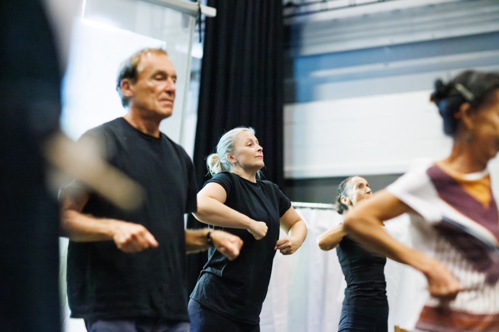 Group of three dancers in black t-shirts stand in a line, bending their arms.