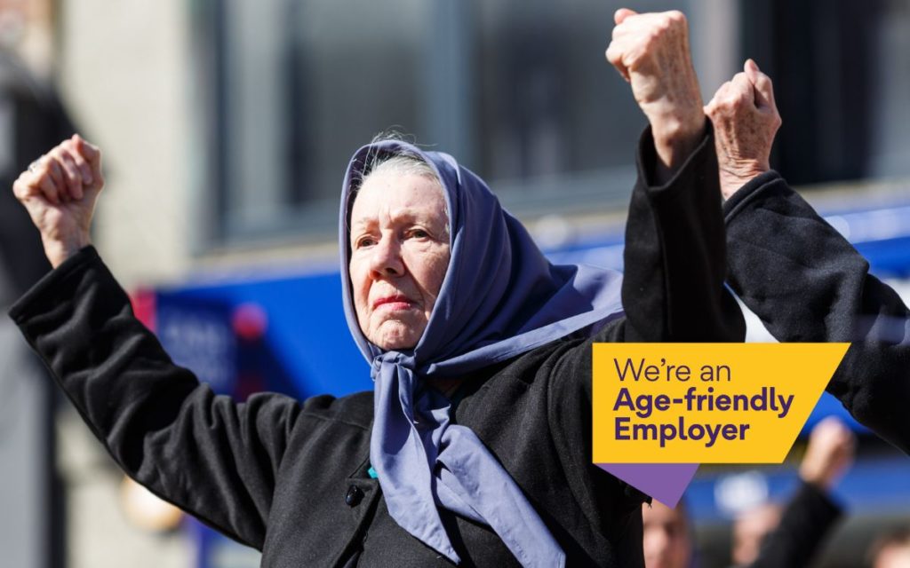 Older woman performer raising her fists confidently in a public outdoor setting, wearing a headscarf and coat. A badge overlay in the corner reads “We’re an Age-friendly Employer.”