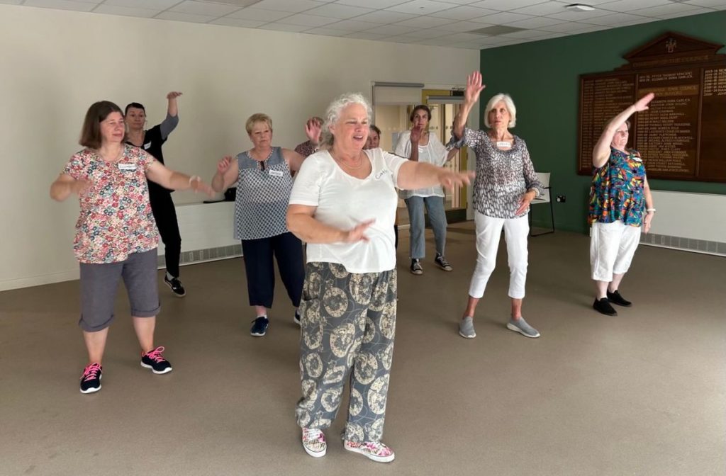A group of older women dance and hold up their arms during a movement workshop.
