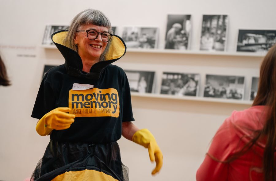 A Moving Memory performer wearing a black t-shirt with the Moving Memory logo and a vibrant yellow-and-black costume with marigold accents, smiling while leading a movement workshop.
