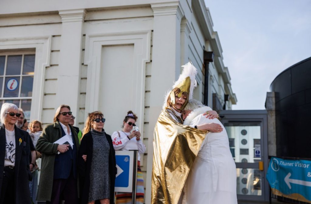 An outdoor performance of Golden Slumbers captures a moment where two women embrace in front of an audience. One wears a gold cape and headdress, hugging the other, who is dressed in a white nightgown.