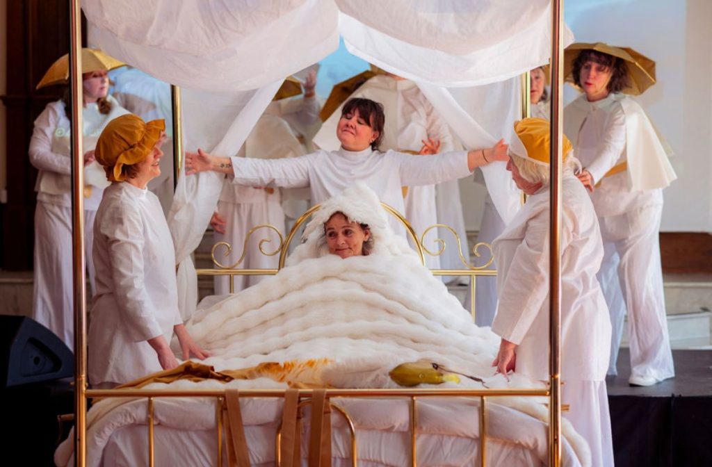 Moving Memory core company perform Golden Slumbers. The group surround a four-poster bed, wearing white. One performer sits on the best, wrapped in a cosy white blanket, smiling.