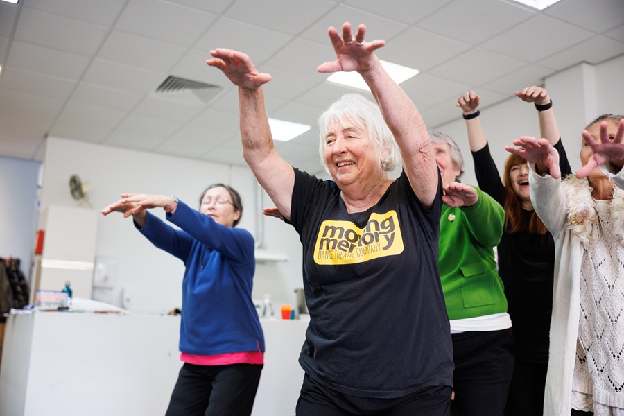 A group of older women dance with their hands held out in front of them. A lady with grey hear stands in the centre, smiling and wearing a black Moving Memory t-shirt.