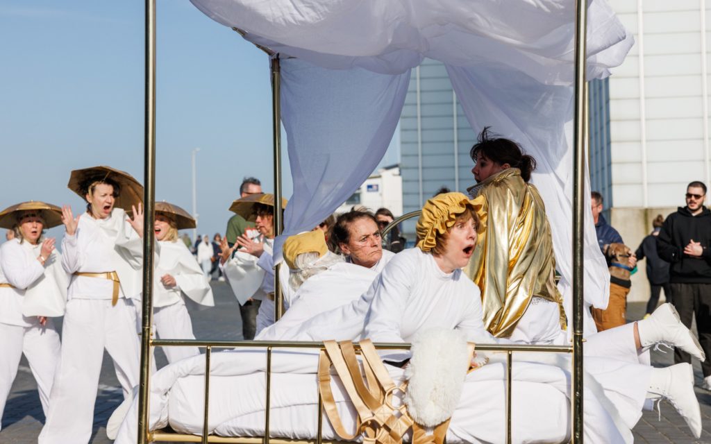 A group of performers in white and gold costumes perform with a golden bed on wheels outdoors.