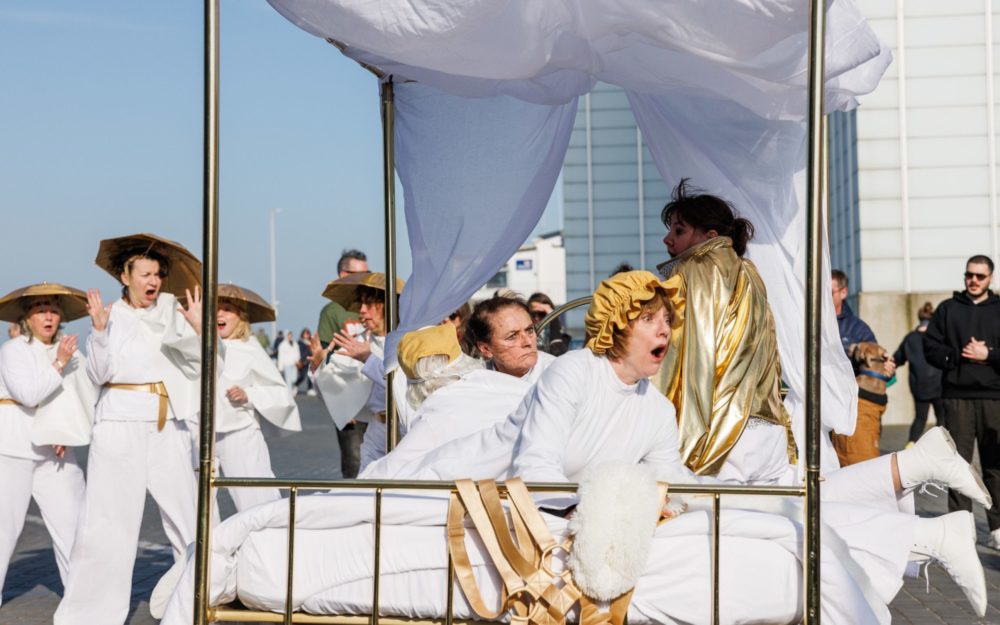 A group of performers in white and gold costumes perform with a golden bed on wheels outdoors.