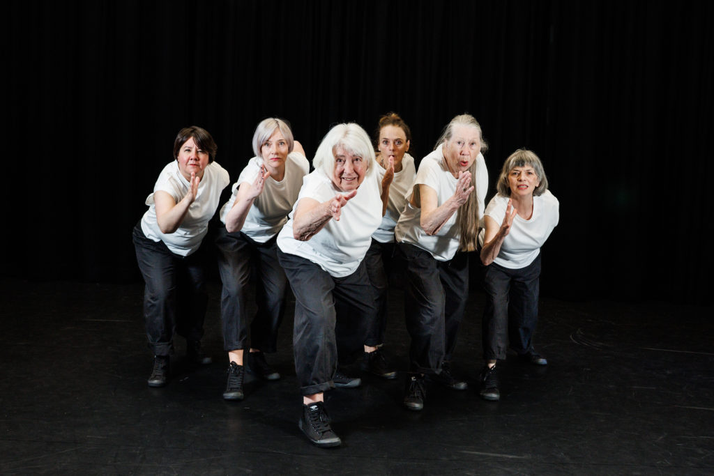 Six older women from Moving Memory’s core company wear white shirts and lean forward in a stylised running pose, lined up side by side.