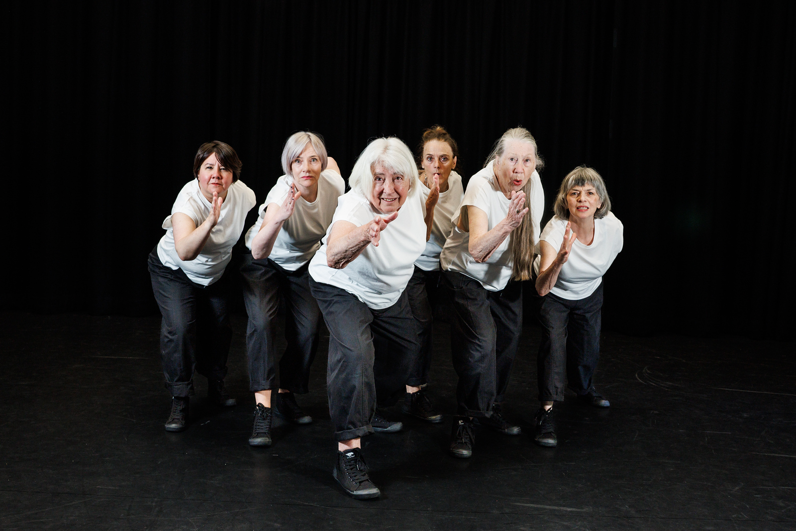 Six older women from Moving Memory’s core company wear white shirts and lean forward in a stylised running pose, lined up side by side.
