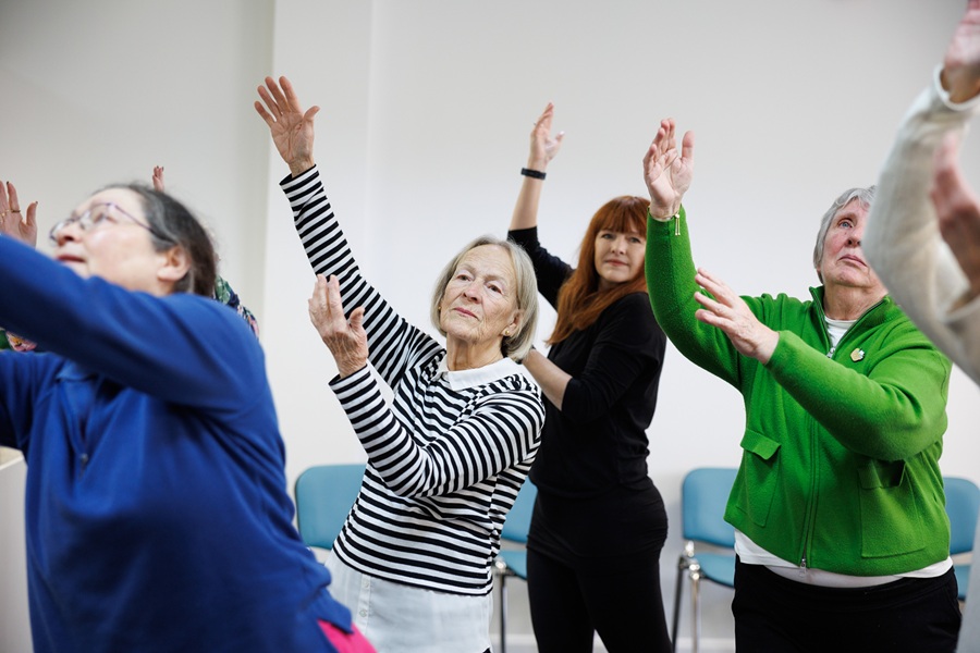 Four older women dance in a line, with their hands reaching over their heads. They wear colourful tops.