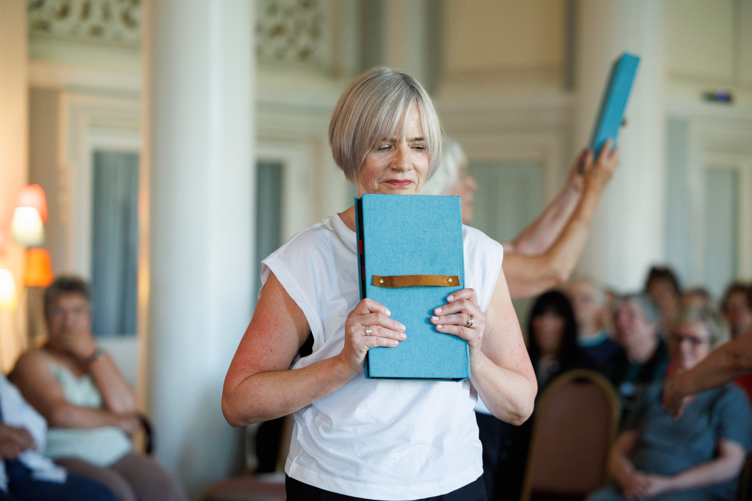 Ginette Casey holds a light blue, rectangular box against her chest with a focused expression during a Moving Memory performance. She wears a white top, with an audience seated softly blurred in the background.