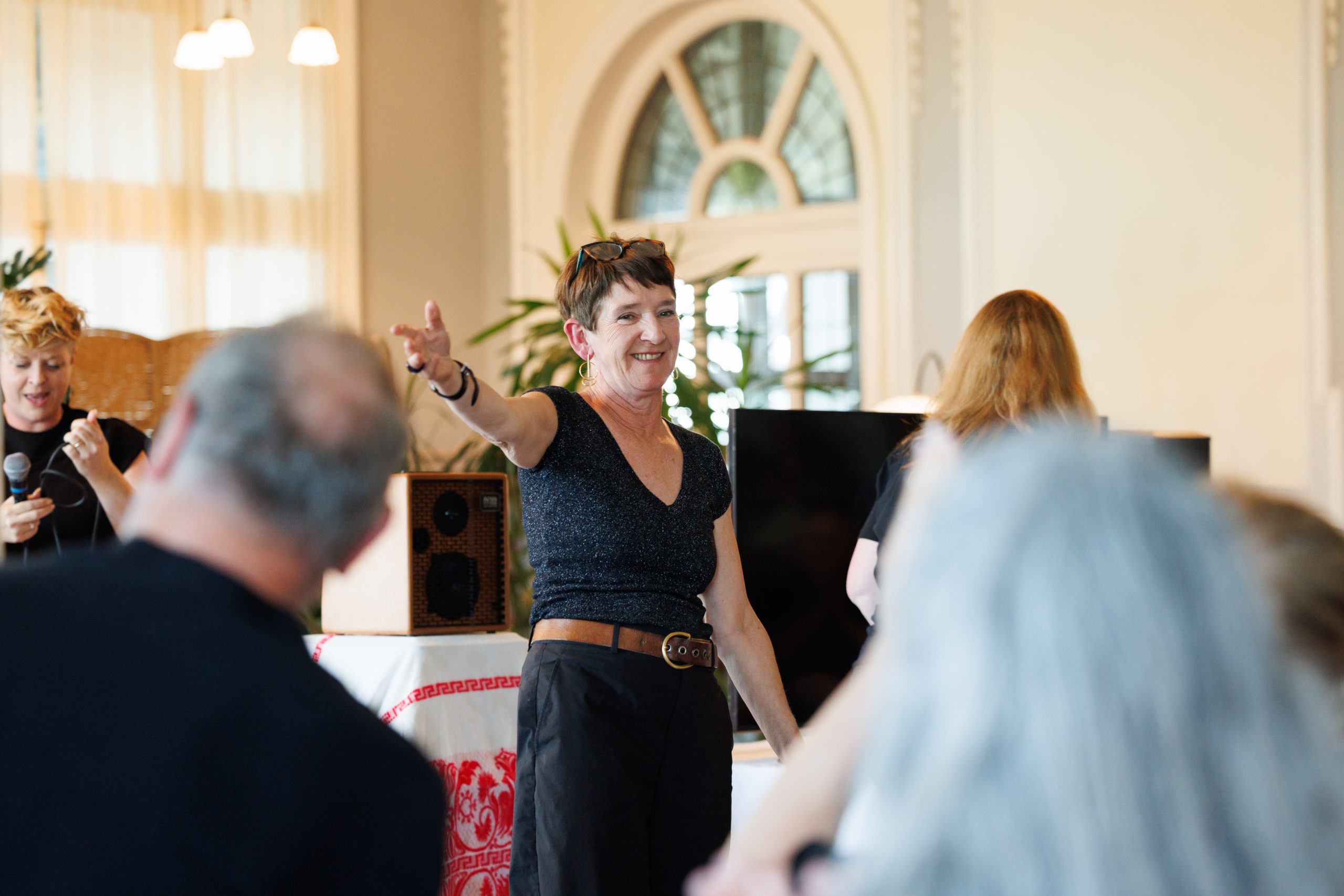 Sian Stevenson stands centre stage in a hall, smiling broadly and gesturing with an outstretched arm toward the audience.
