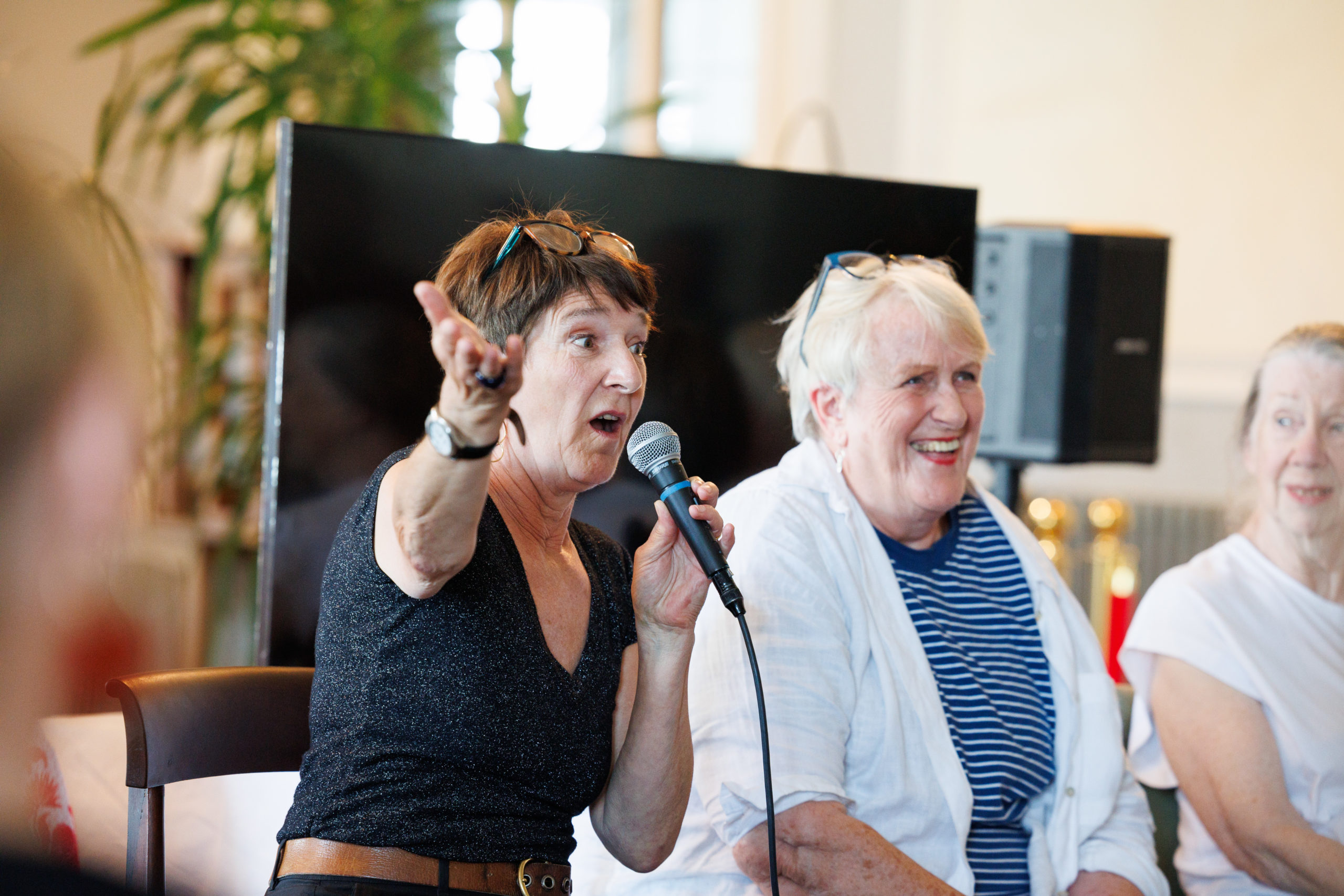 A medium shot of Sian Stevenson speaking animatedly into a microphone with one hand raised; Bobby Baker smiles widely to her right, wearing a blue and white striped shirt.
