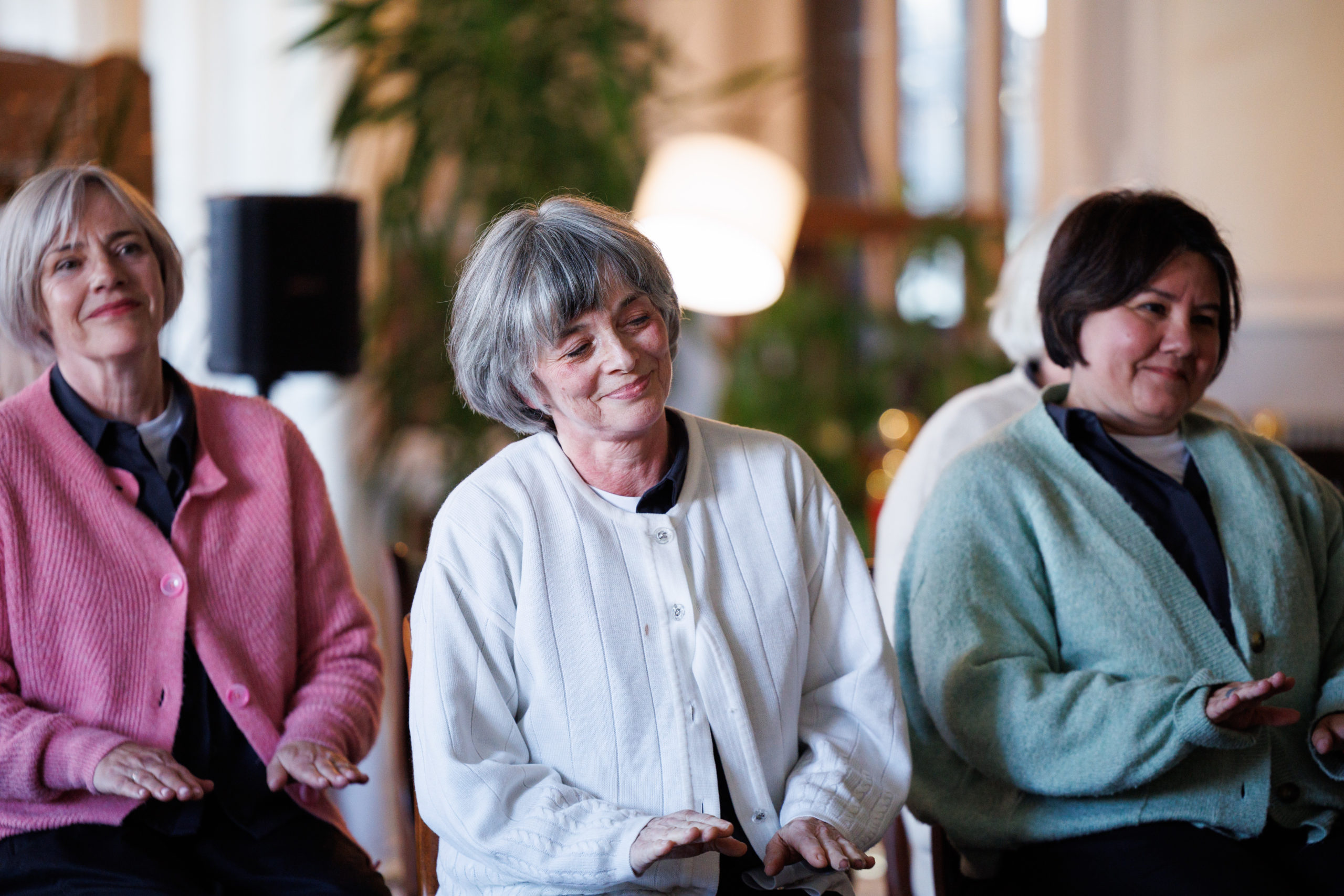 Three seated Moving Memory performers look on with subtle, gentle smiles during a quiet moment of the performance, wearing pink, white, and sage green cardigans.