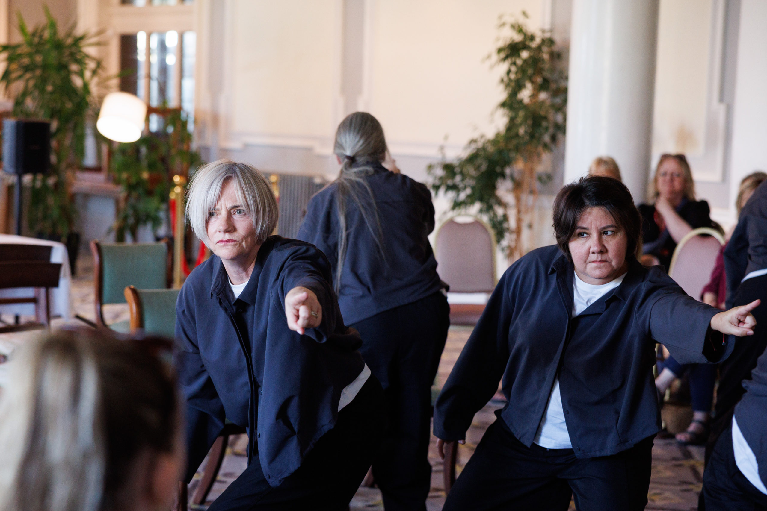 Performers Ginette Casey and Meire Machado lean forward in a crouched performance stance, pointing sharply outwards with serious, focused expressions. Both wear dark grey button-up shirts over white tops.