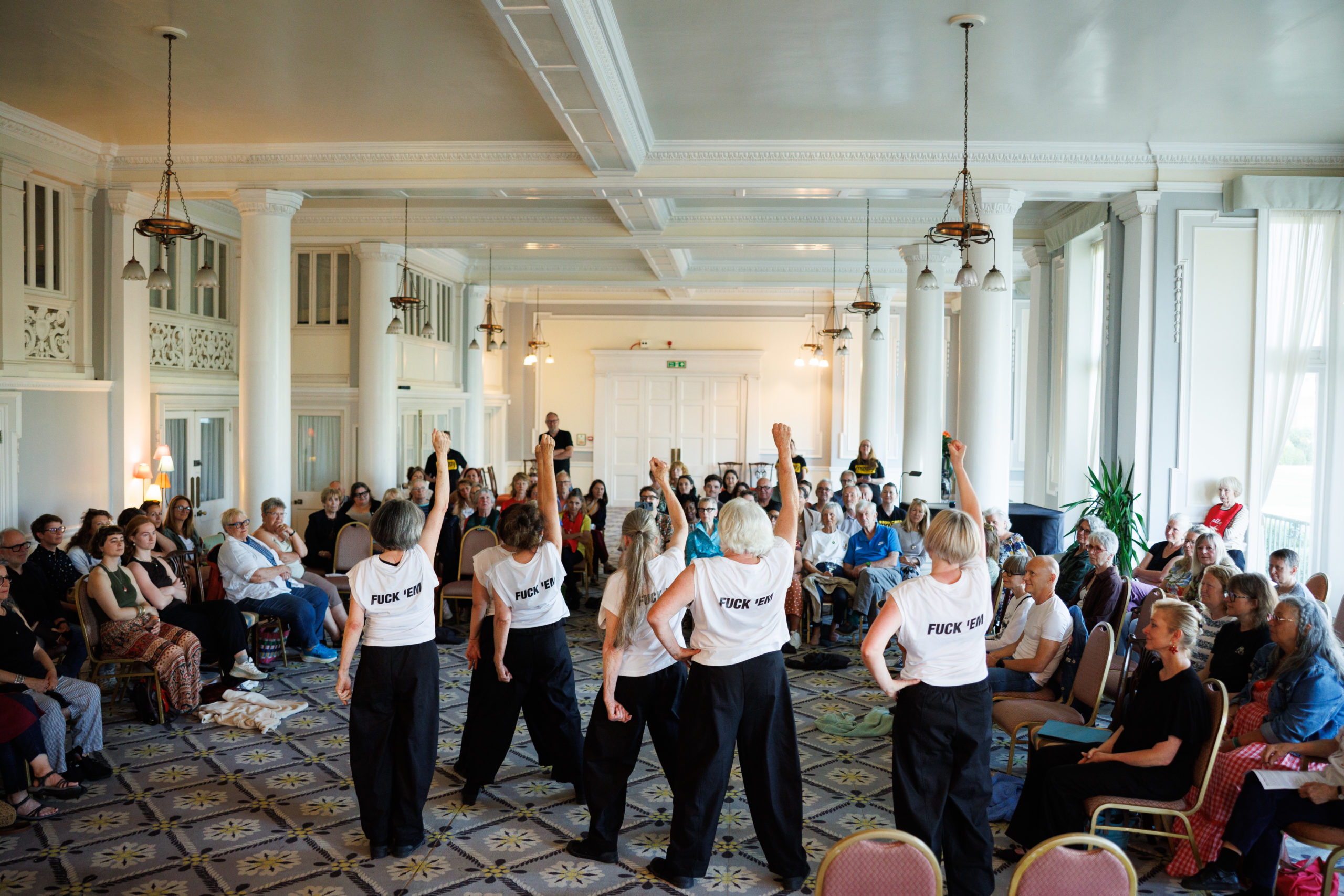Moving Memory performers stand in a circle facing an audience, raising their fists in solidarity. The back of their white t-shirts reads "FUCK 'EM".