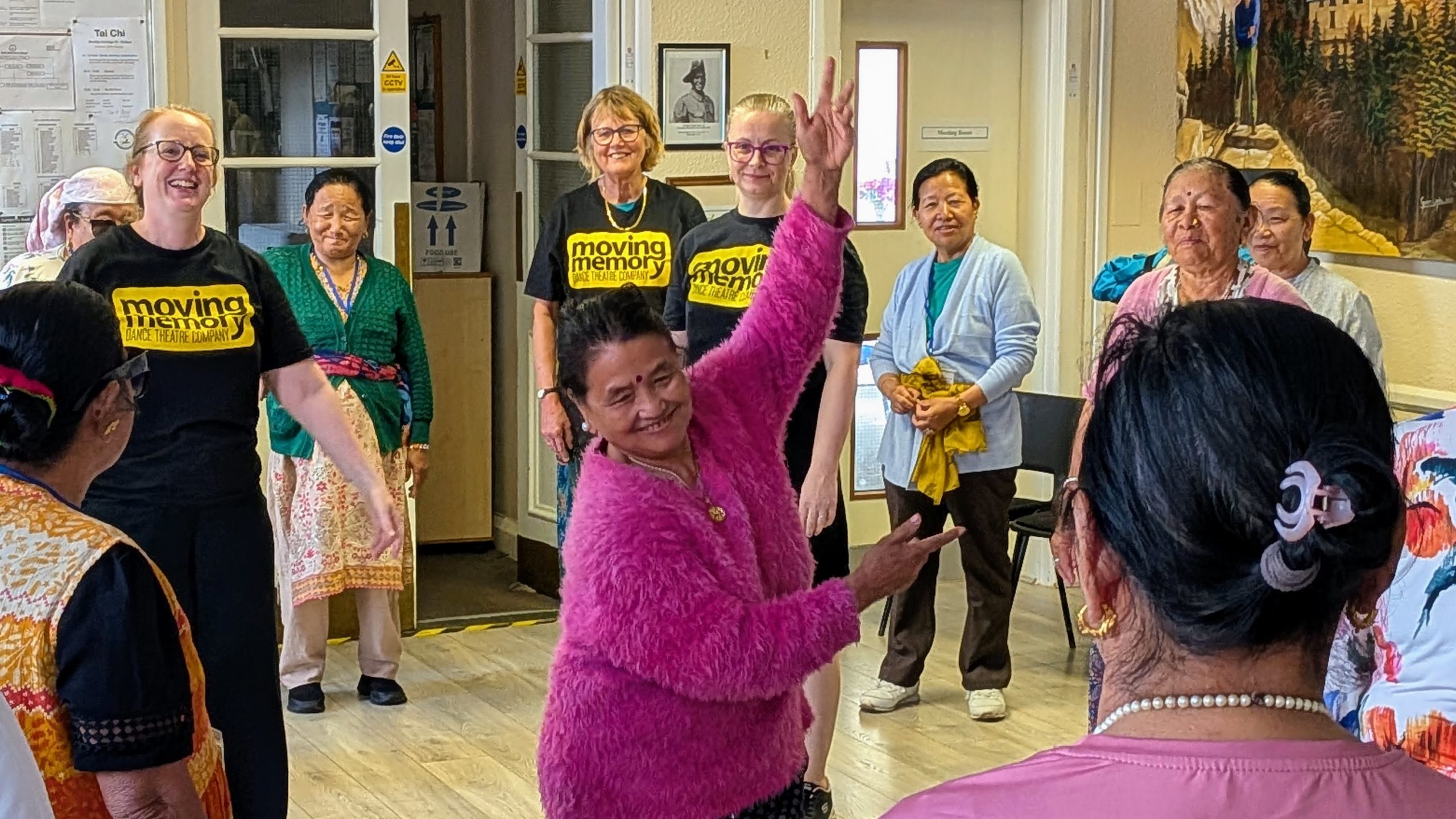 A woman in a bright pink fluffy cardigan dances joyfully at the centre of a circle, raising one arm high while Moving Memory facilitators and group members watch with smiles.