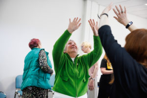 smiling woman in a bright green fleece reaches both hands high into the air, mirroring another participant during an indoor workshop exercise.