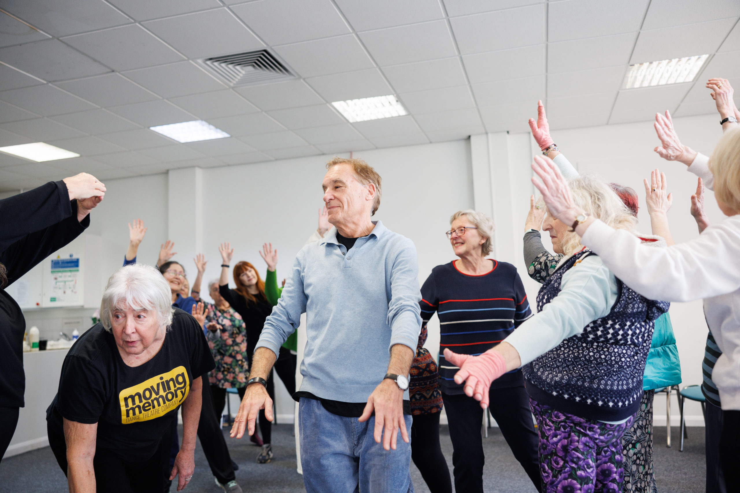 A group of older participants practice movement together in a bright studio, guided by a workshop leader leaning forward in a Moving Memory t-shirt.